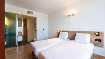 Habitación de hotel con dos camas, cabecero de madera, luces fijadas a la pared y puerta corredera hacia un baño.
