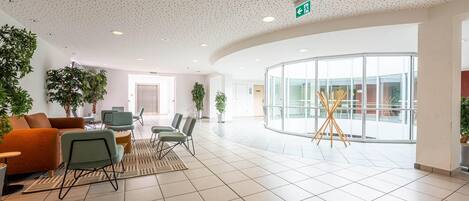 A spacious lobby with a high ceiling, a seating area with chairs and sofas, a large glass wall, and potted plants.
