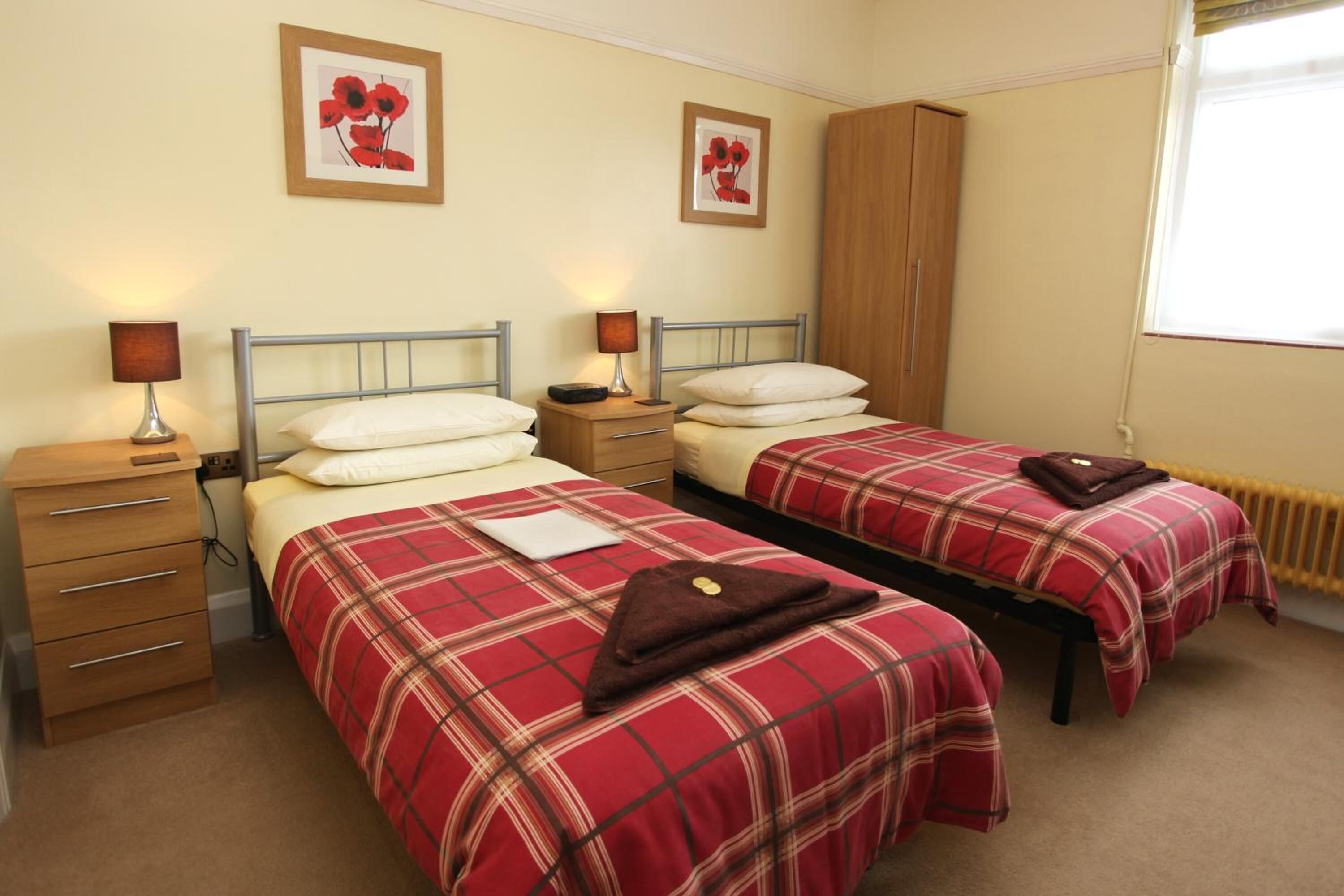 A twin room with two beds, each with a red plaid bedspread, bedside tables with lamps, a wooden wardrobe, and framed artwork on the walls.