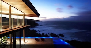 A modern seaside property with a glass balcony overlooking the ocean, a wooden deck, and a pool with blue lighting. - 3 The Cove (Knysna)