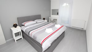 Ein modernes Schlafzimmer mit grauem Bett, weißen Nachttischen, einem Schreibtisch und einem großen Fenster mit leichtem Vorhang.