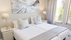 Un lit double avec du linge de lit blanc, deux tables de chevet avec des lampes, un plaid à motifs et un grand tableau abstrait accroché au mur.