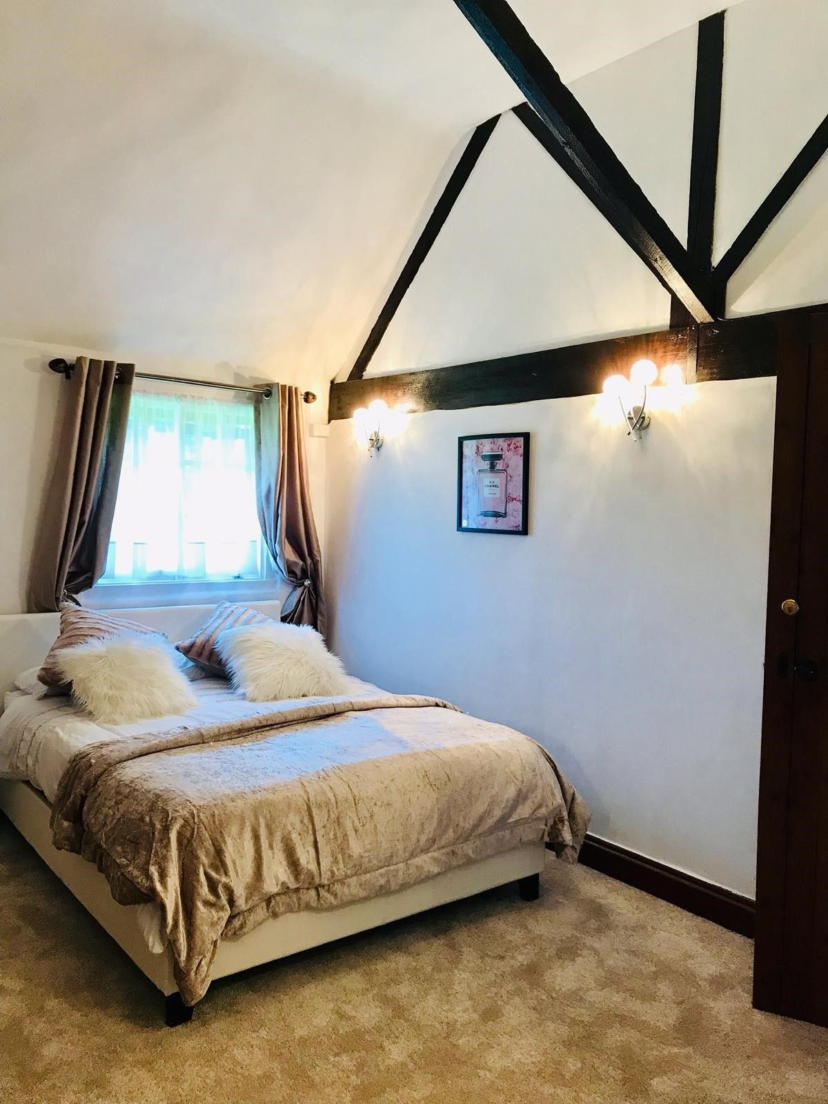 A bedroom with a bed, a window with curtains, a wall-mounted light fixture, and a framed picture.