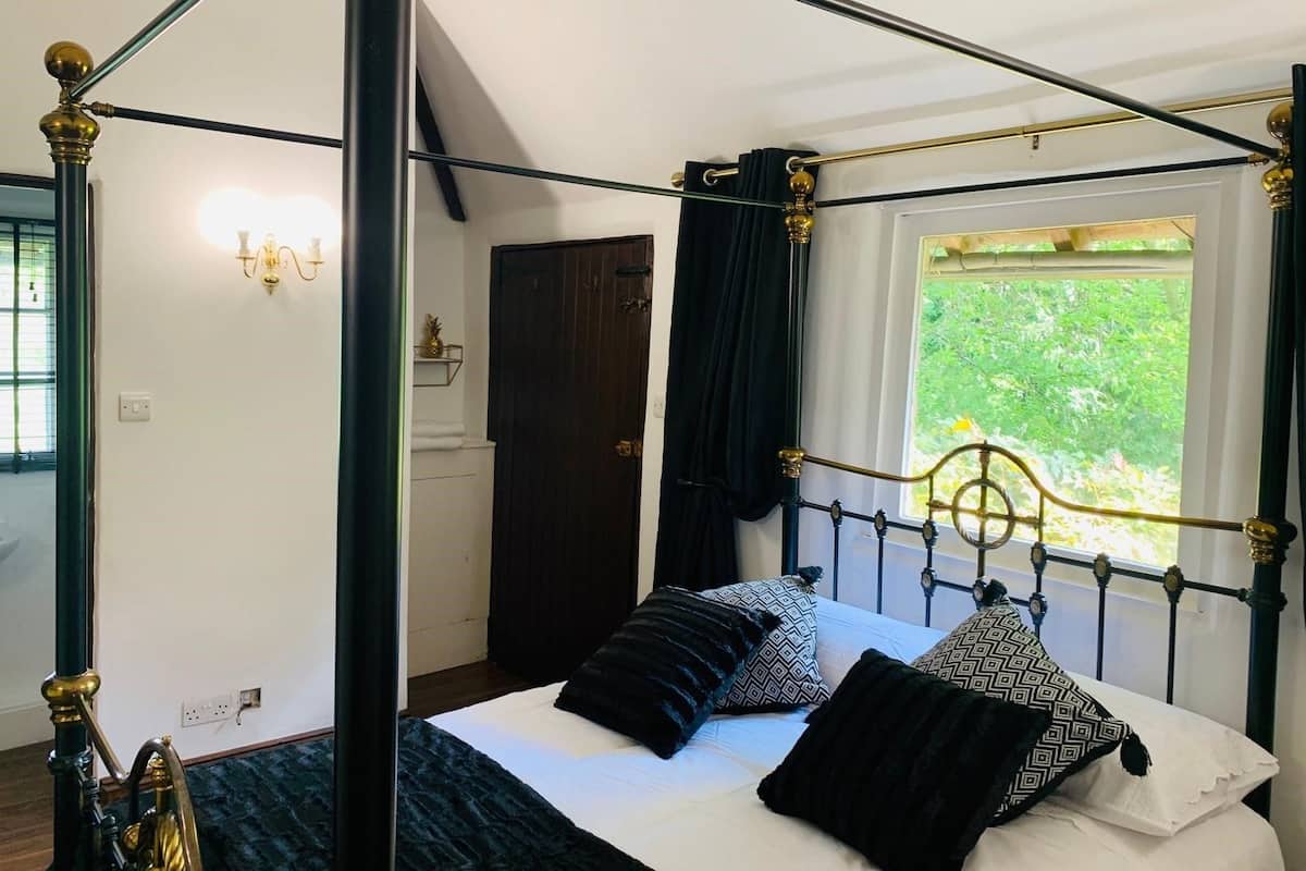 Ein Bett mit Baldachin, mit dunklem Metallgestell und goldenen Akzenten, einer weißen Bettdecke und gemusterten Kissen. Ein Fenster bietet Blick auf Grünflächen im Freien.