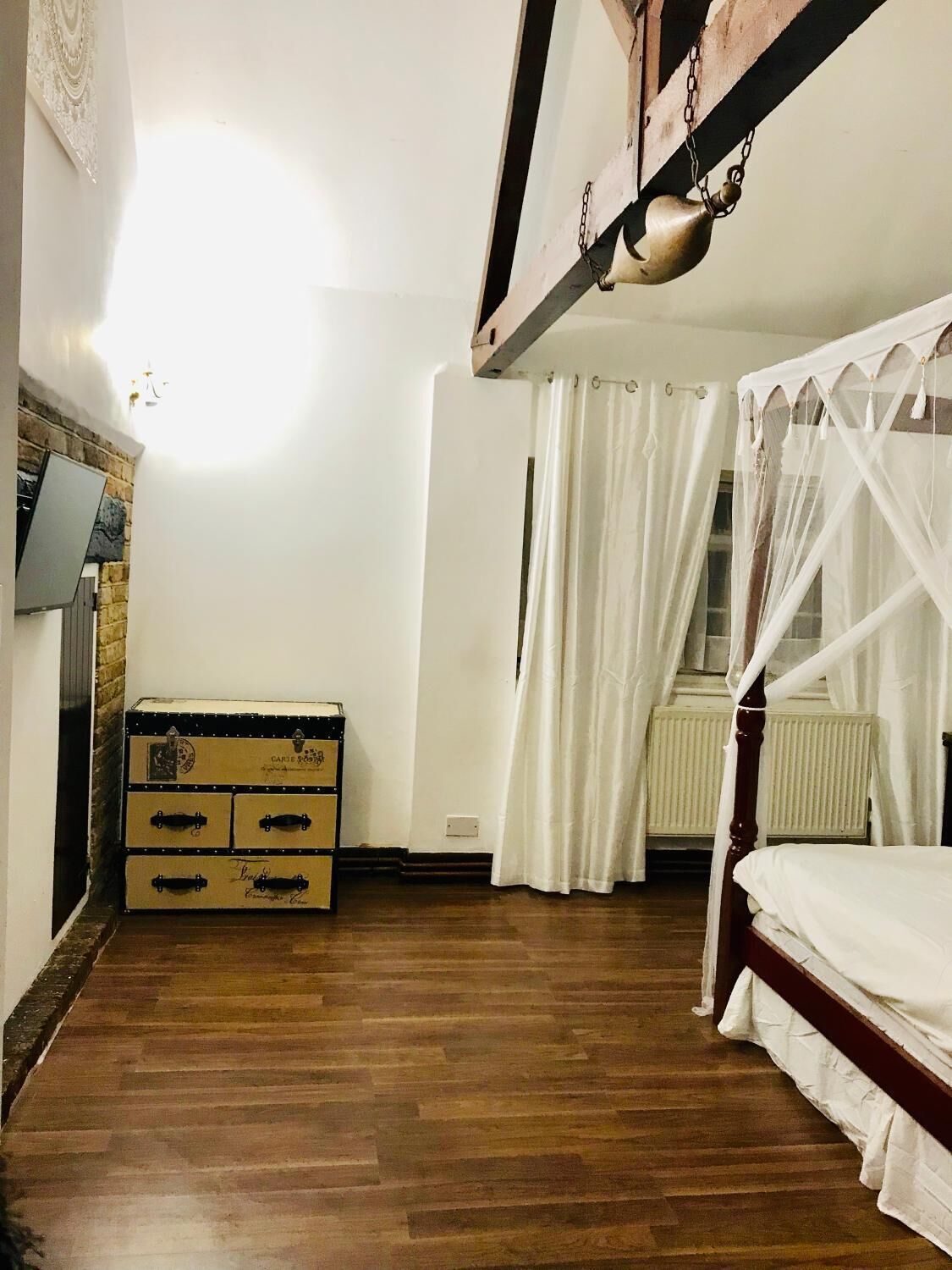 Un dormitorio con suelo de madera, cama con dosel blanco, cómoda, radiador y televisor montado en la pared.
