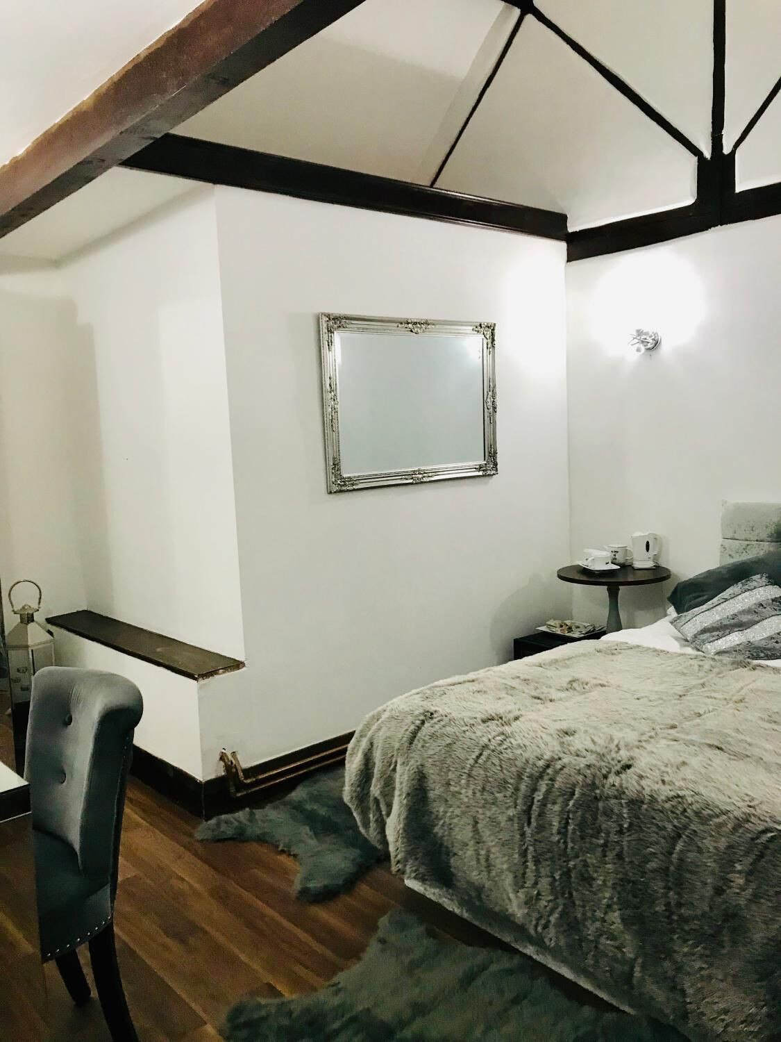 A bedroom with a bed, a chair, a small table with a lamp, and a mirror on the wall.