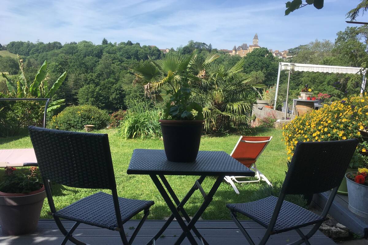 Un patio con dos sillas y una mesa, rodeado de exuberante follaje y una vista a un castillo a lo lejos.