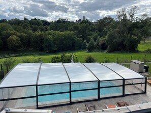 Una estructura tipo invernadero con una piscina debajo, rodeada de un jardín y árboles.