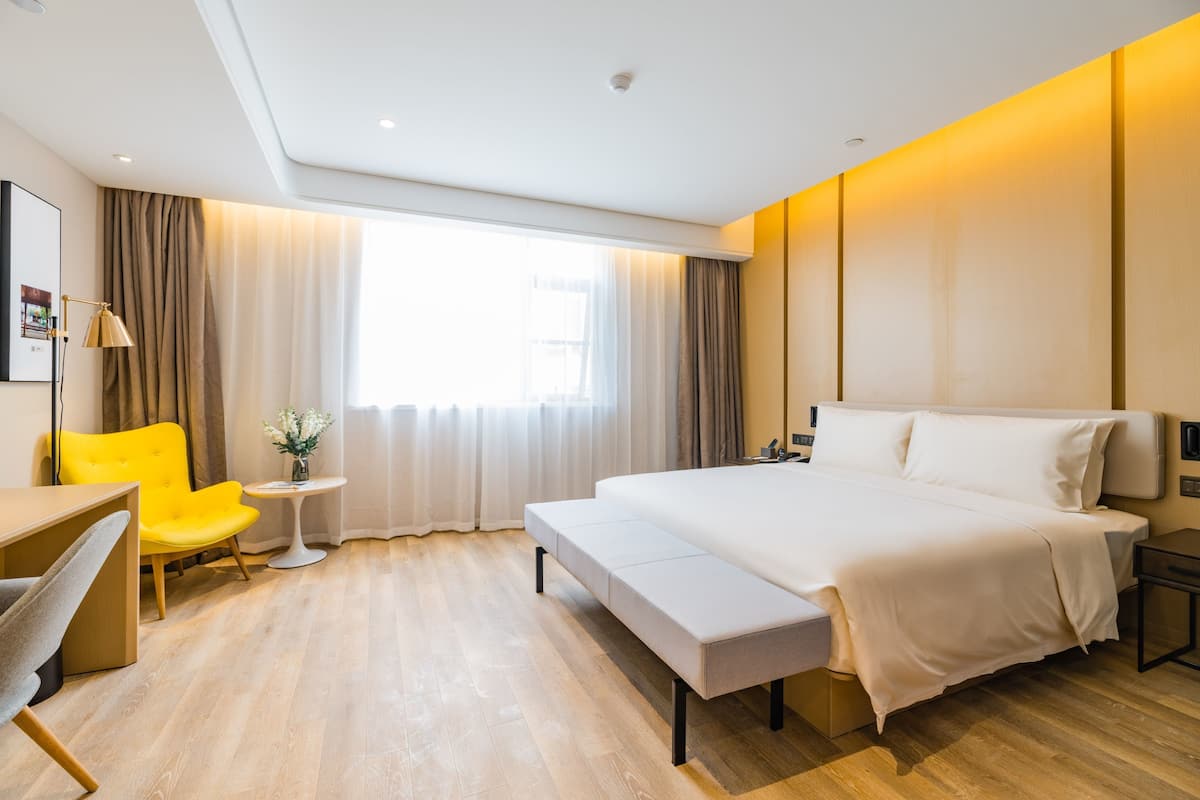 Ein modernes Hotelzimmer mit einem großen Bett, einem Schreibtisch mit Stuhl, einem kleinen Tisch mit Blumenvase und einem großen Fenster mit Vorhängen.