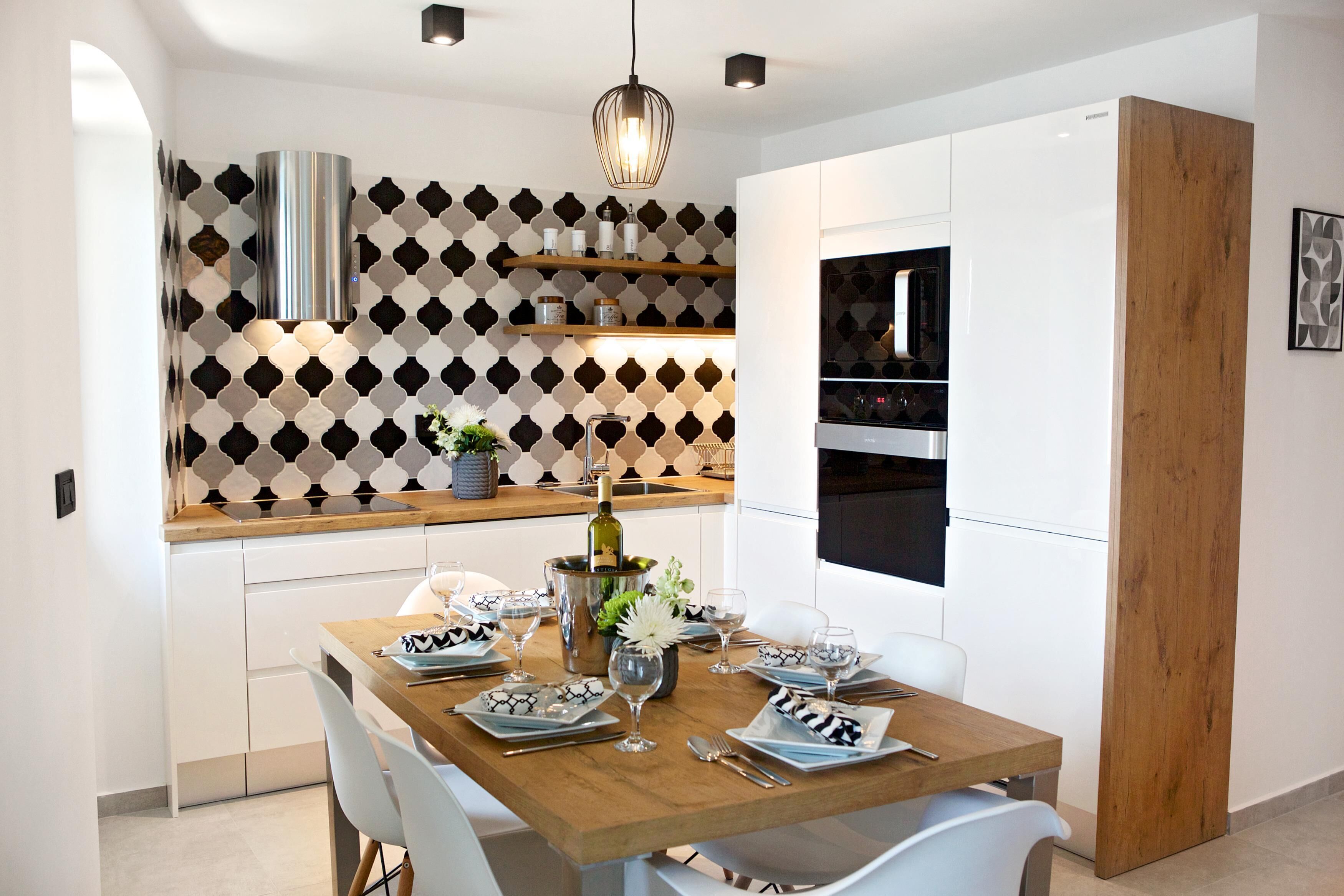 A modern kitchen with white cabinets, a wooden countertop, and a patterned backsplash. There is a dining table set with plates, glasses, and a bottle of champagne.