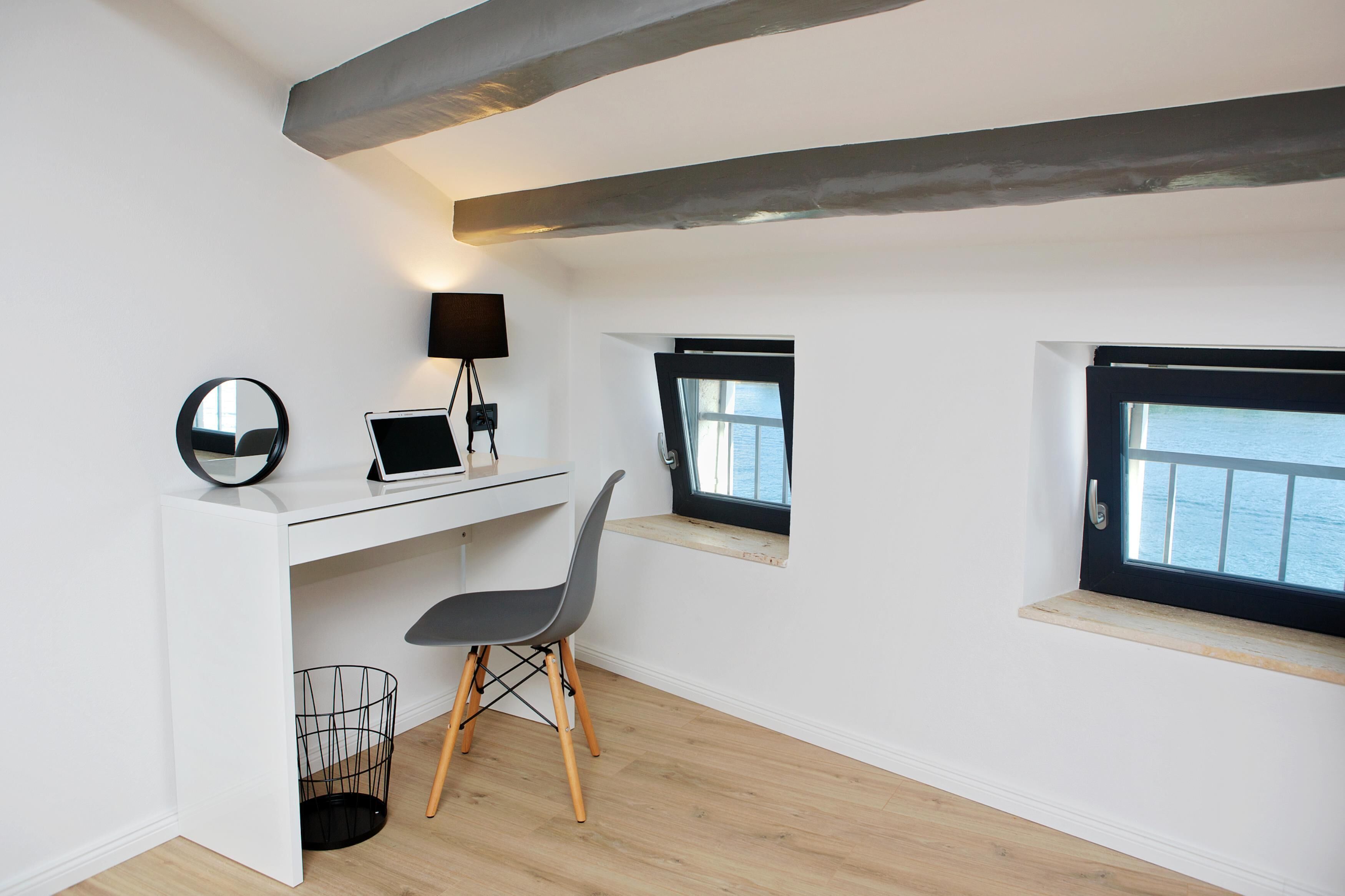 A modern, minimalist room with a white desk, a laptop, a chair, and a lamp. There are two windows with views of the sea.