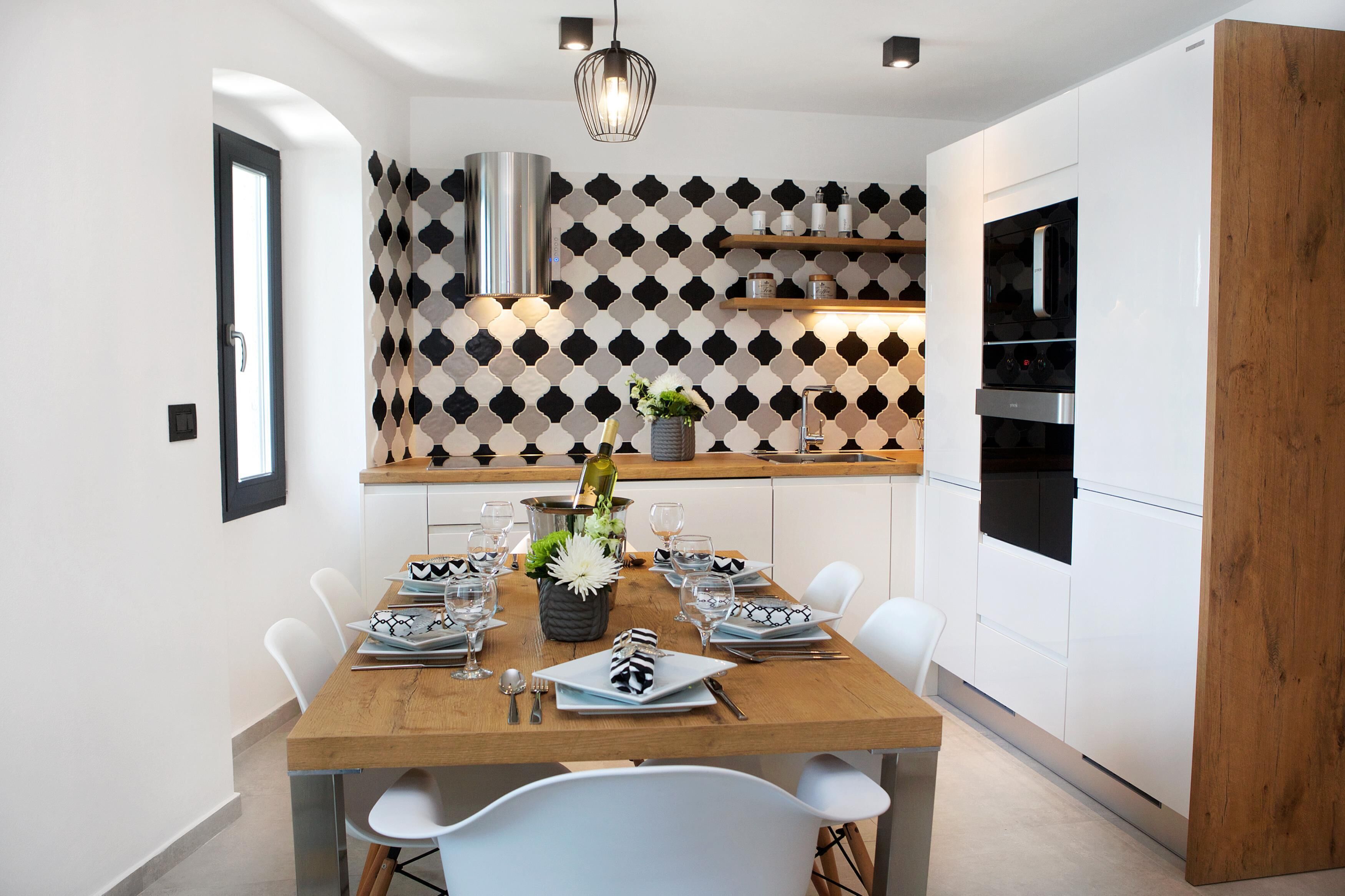 A modern kitchen with white cabinets, a wooden dining table set for six, and a patterned backsplash.