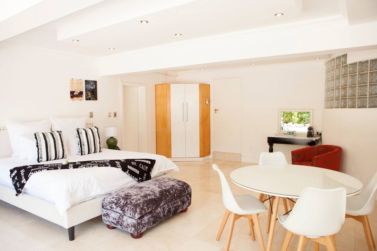 A modern bedroom with a bed, bedside table, and a dining area with chairs.