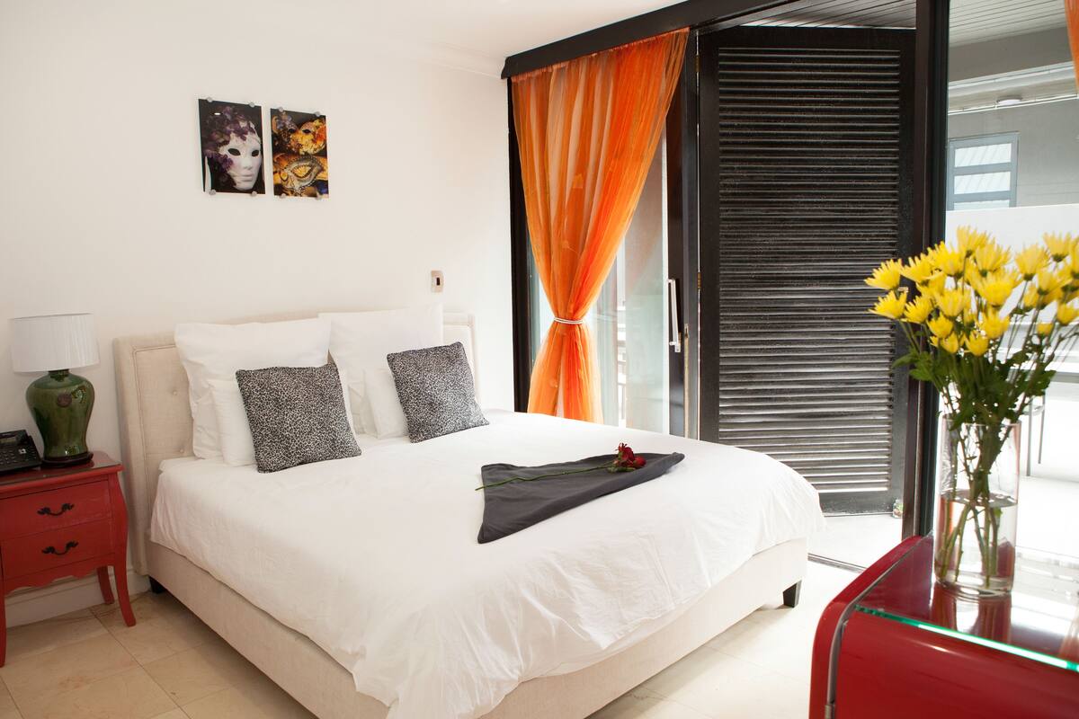A neatly made bed with white linens, a red nightstand with a green vase, and a vase of yellow flowers on a glass table.