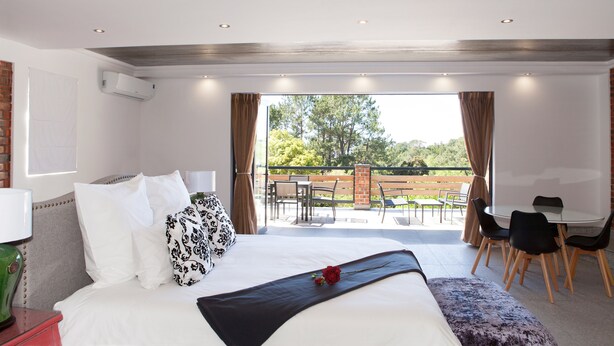 A modern hotel room with a large bed, a balcony with outdoor seating, and a view of the outdoors.
