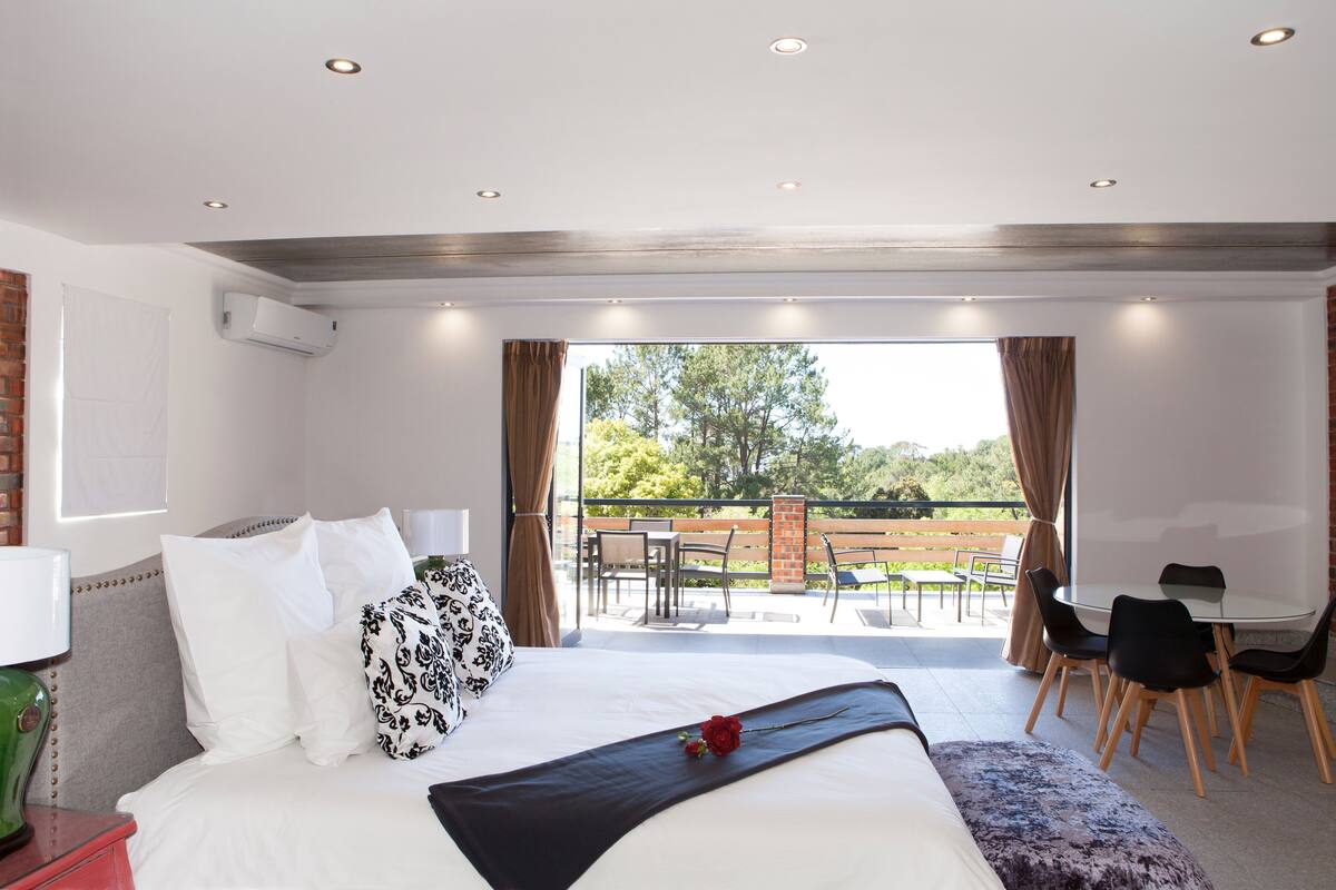 A modern hotel room with a large bed, a balcony with outdoor seating, and a view of the outdoors.