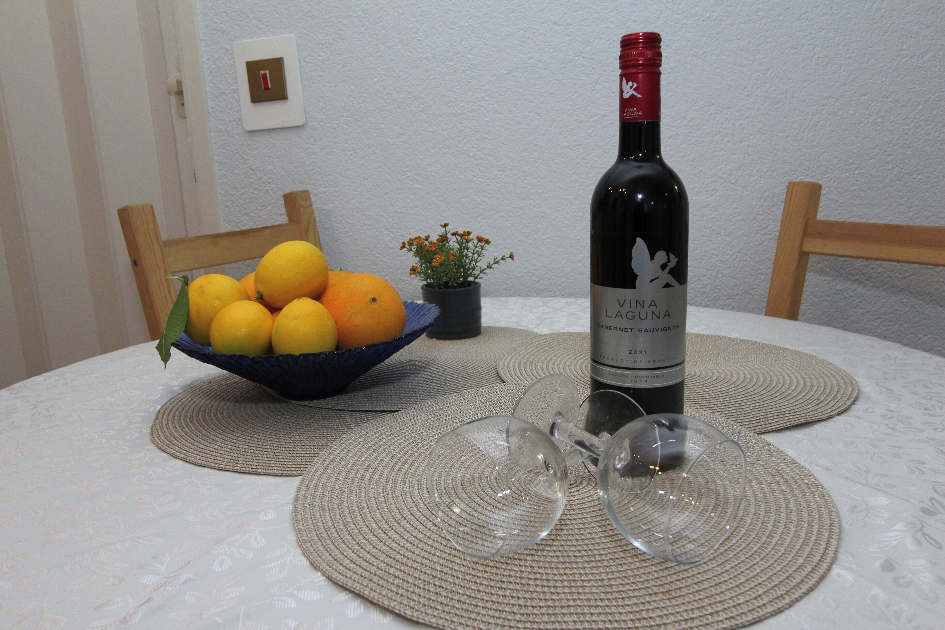 A table with a bottle of Vina Cacumaga wine, two empty wine glasses, a bowl of lemons and oranges, and a small potted plant.