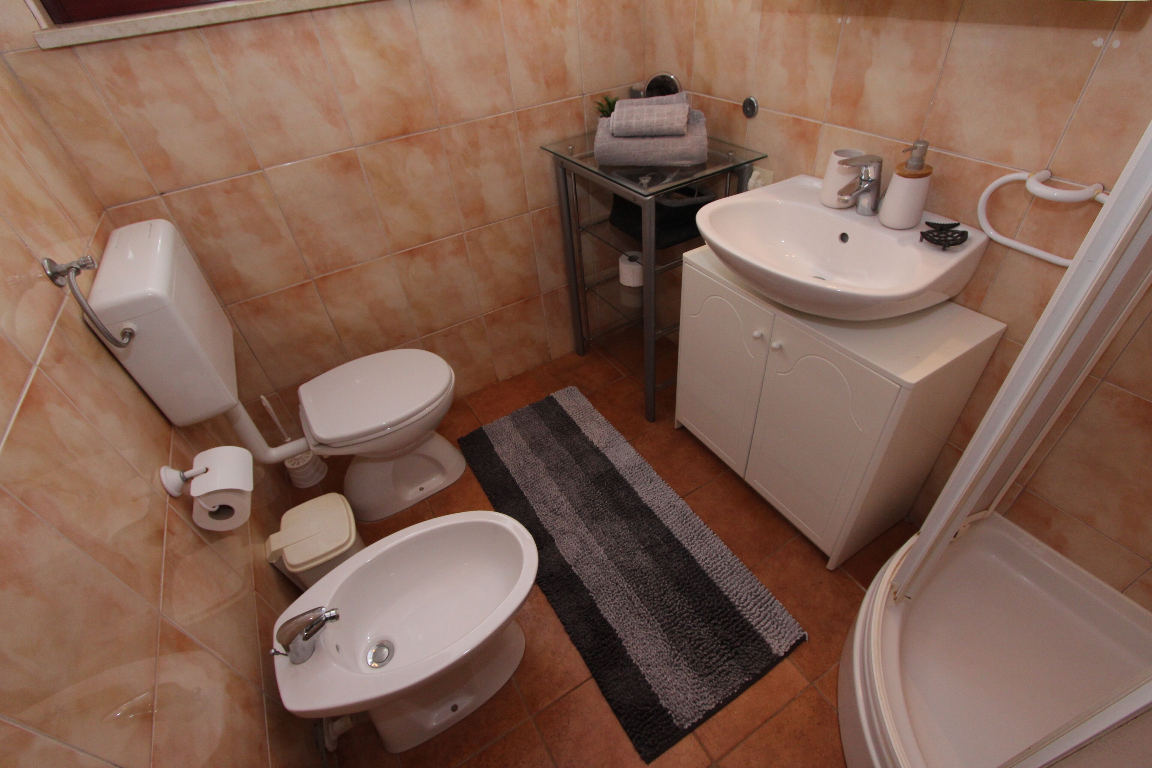 Casa de banho compacta com sanita, bidé, lavatório e uma pequena mesa de vidro com produtos de higiene pessoal.