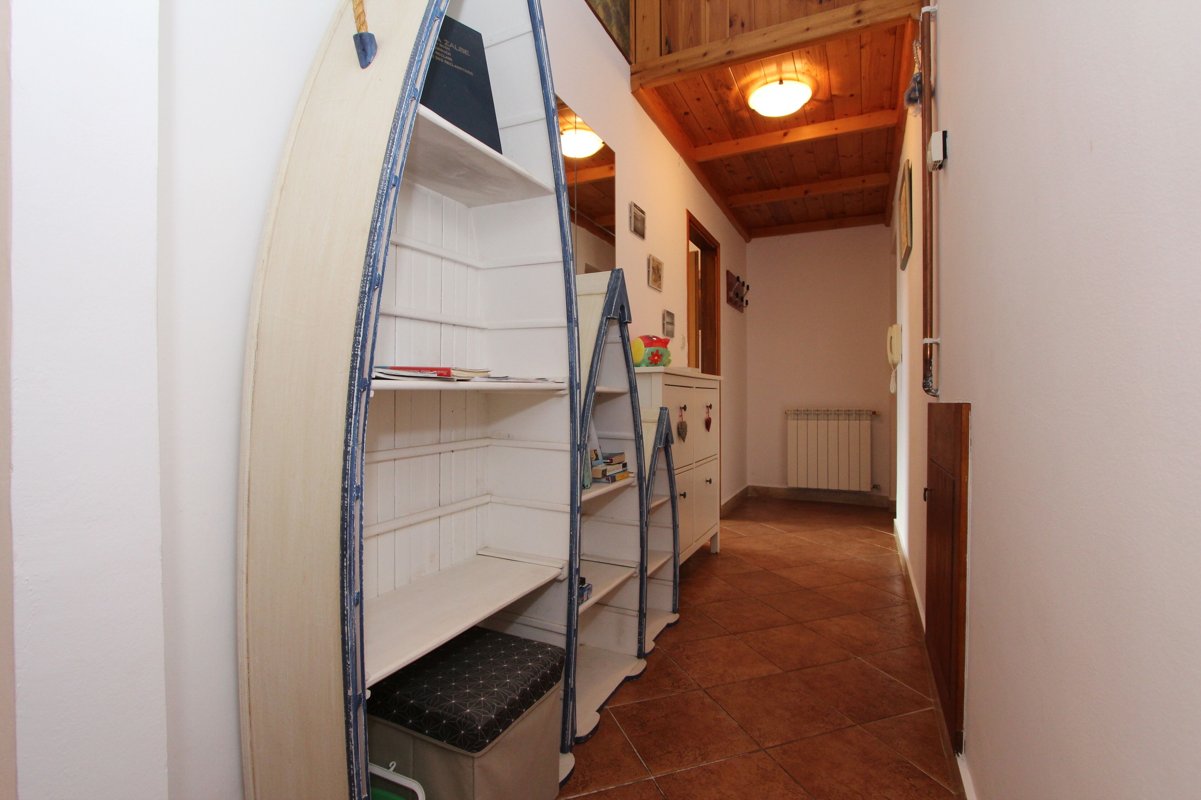A narrow hallway with wooden beams on the ceiling and a radiator on the wall. Shelves and a ladder are visible in the room.