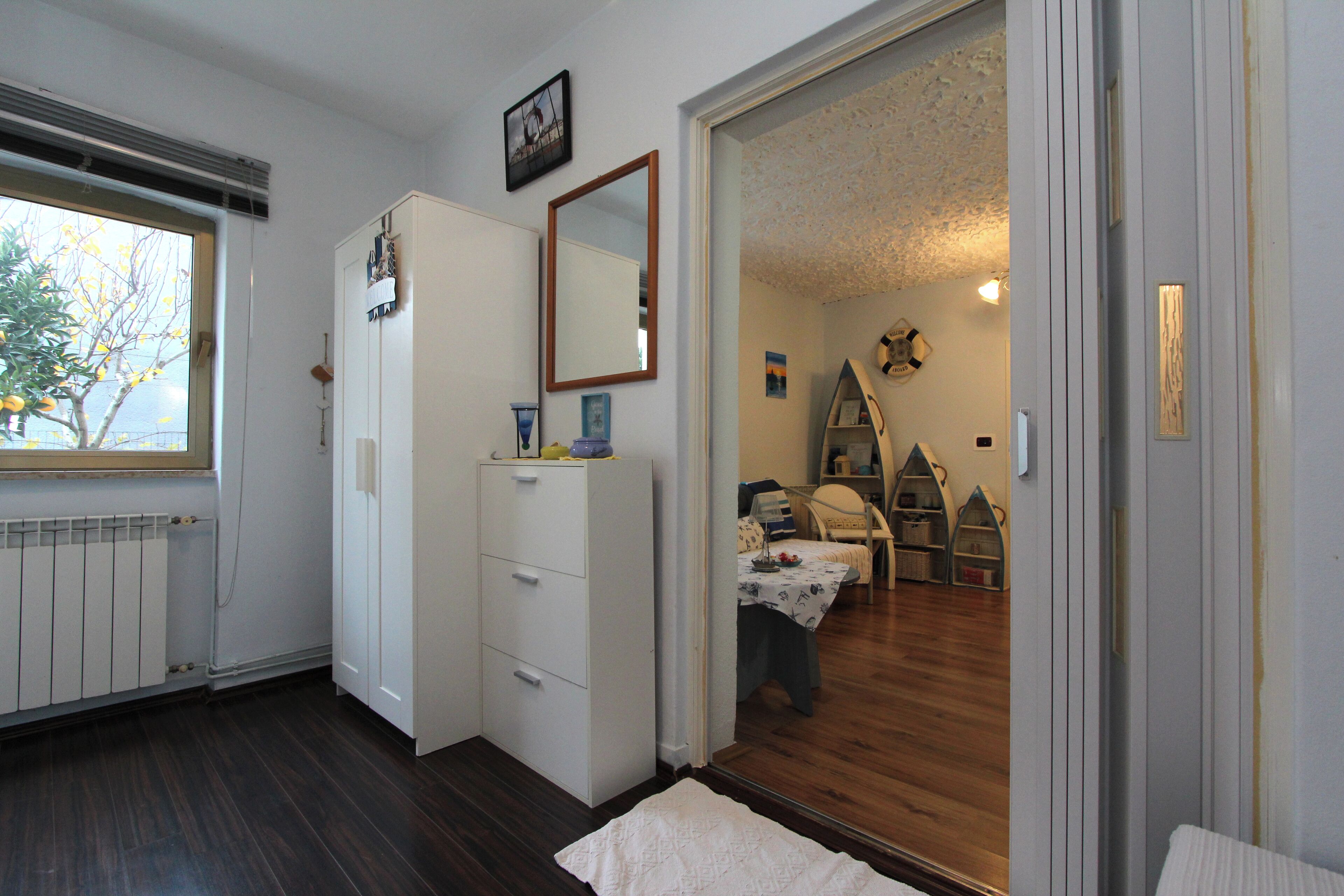 A room with a radiator, a white wardrobe, a mirror, and a view of another room with a bed and a wooden floor.