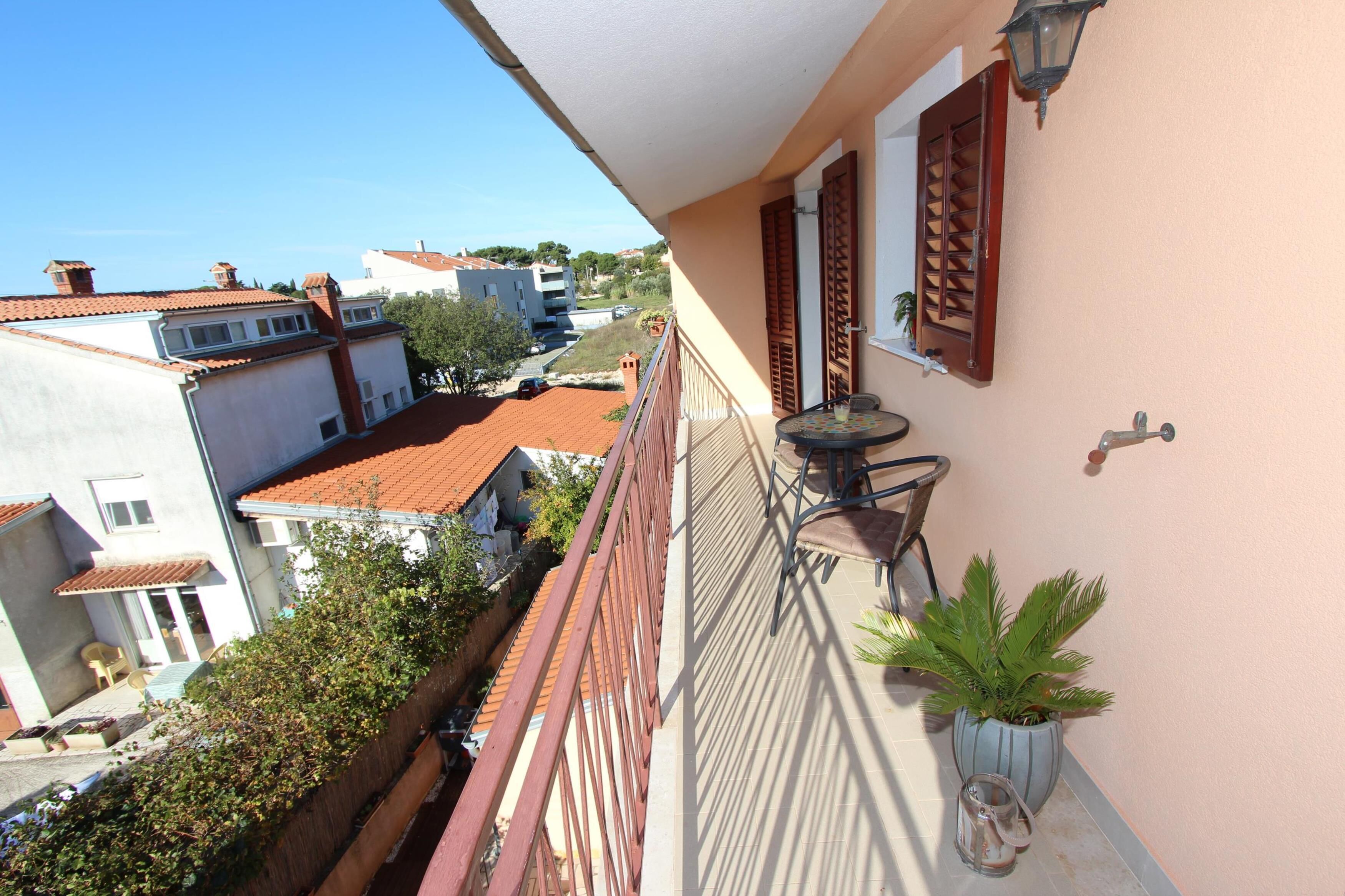 A balcony with a view of neighboring buildings, a potted plant, and a pair of shoes on the floor.