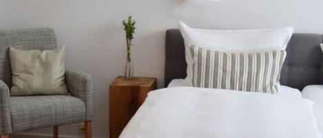 A neatly made bed with white bedding, a grey upholstered armchair with a cream pillow, and a small wooden nightstand with a vase of green plants.