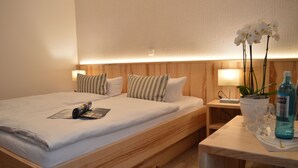 Een keurig opgemaakt bed met witte lakens en gestreepte kussens, een nachtkastje met een lamp, een kleine potplant en een fles water.