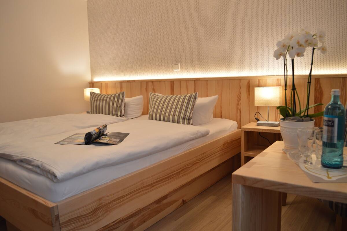 En pænt redt seng med hvide sengetæpper og stribede puder, et natbord med en lampe, en lille potteplante og en vandflaske.