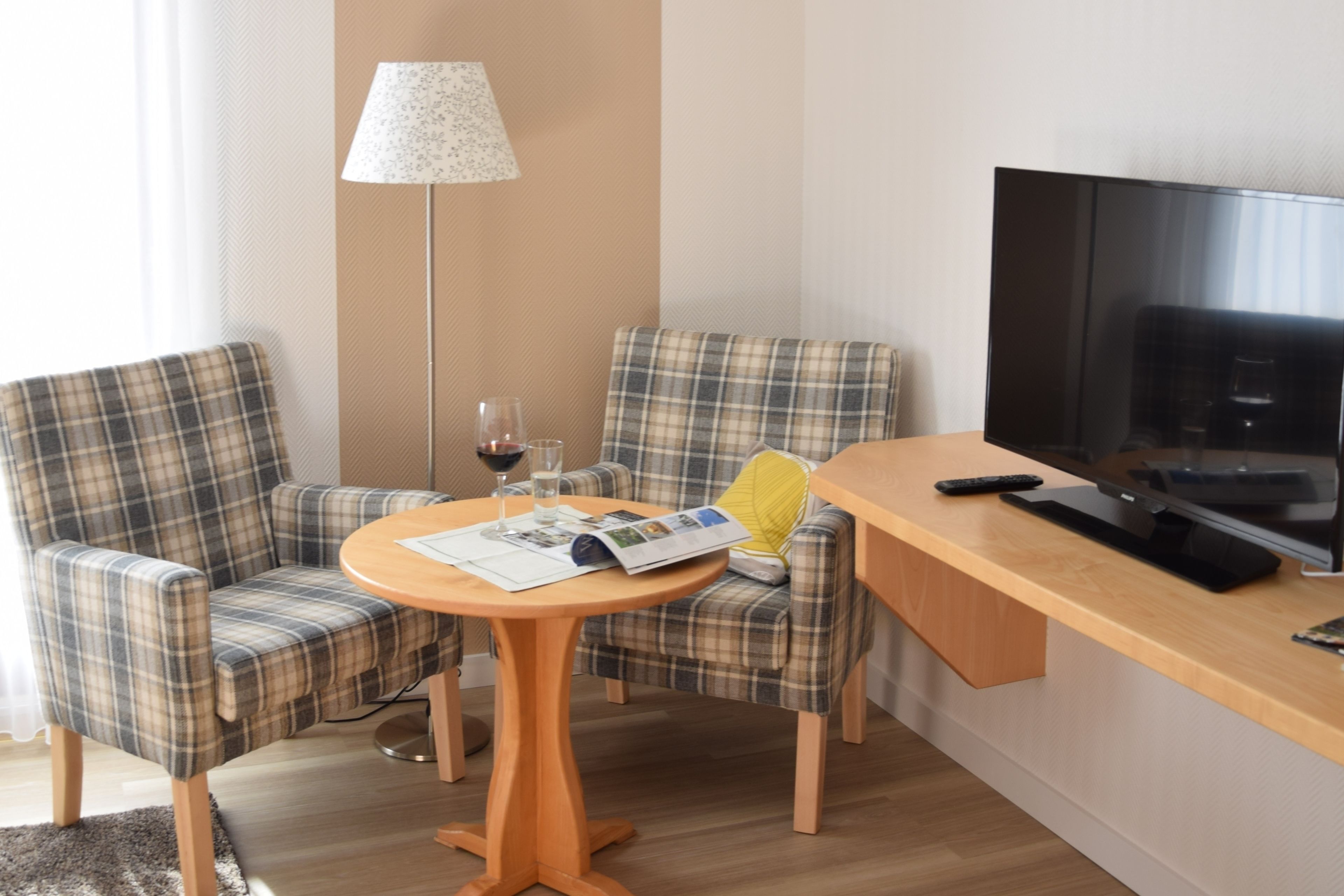 Ein Zimmer mit zwei Karo-Sesseln, einem Holztisch mit einer Zeitschrift, einer Lampe und einem Fernseher auf einem Holzständer.