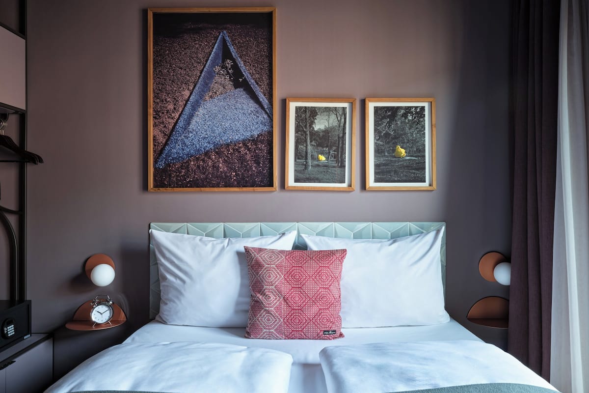 A neatly made bed with white linens and a teal throw blanket, a geometric-patterned pillow, and a wall clock on the nightstand.
