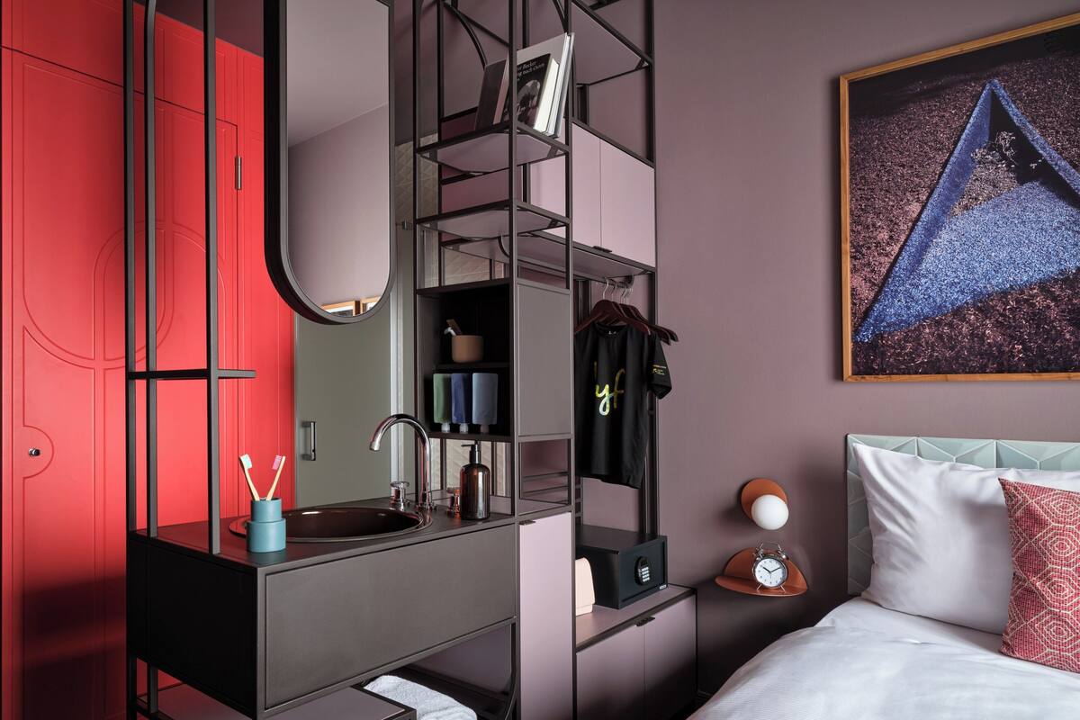 A modern bedroom with a red wardrobe, a built-in vanity with a mirror, a bedside table with a lamp, and a bed with a patterned pillow.