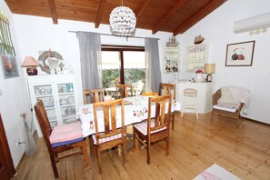 A dining area with a wooden table, chairs, and a chandelier. There is a window with curtains, a bookshelf, and a small stool.