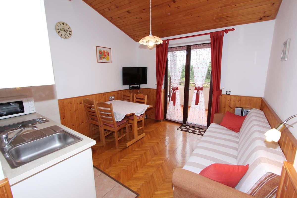 Un cuarto acogedor con techo de madera, una zona de comedor con mesa y sillas, un sofá con cojines rojos, un televisor y una ventana con cortinas.