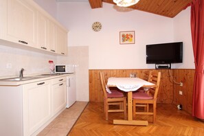 A kitchen with white cabinets, a microwave, and a small refrigerator. There is a dining table with chairs and a TV mounted on the wall. - Apartments Aurora (Rovinj)