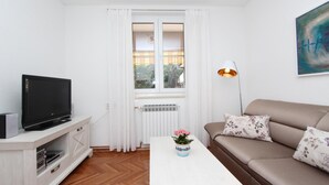Una sala de estar con una televisión sobre un mueble blanco, un sofá con cojines estampados, una mesa de centro blanca con un florero y una ventana con cortinas blancas.