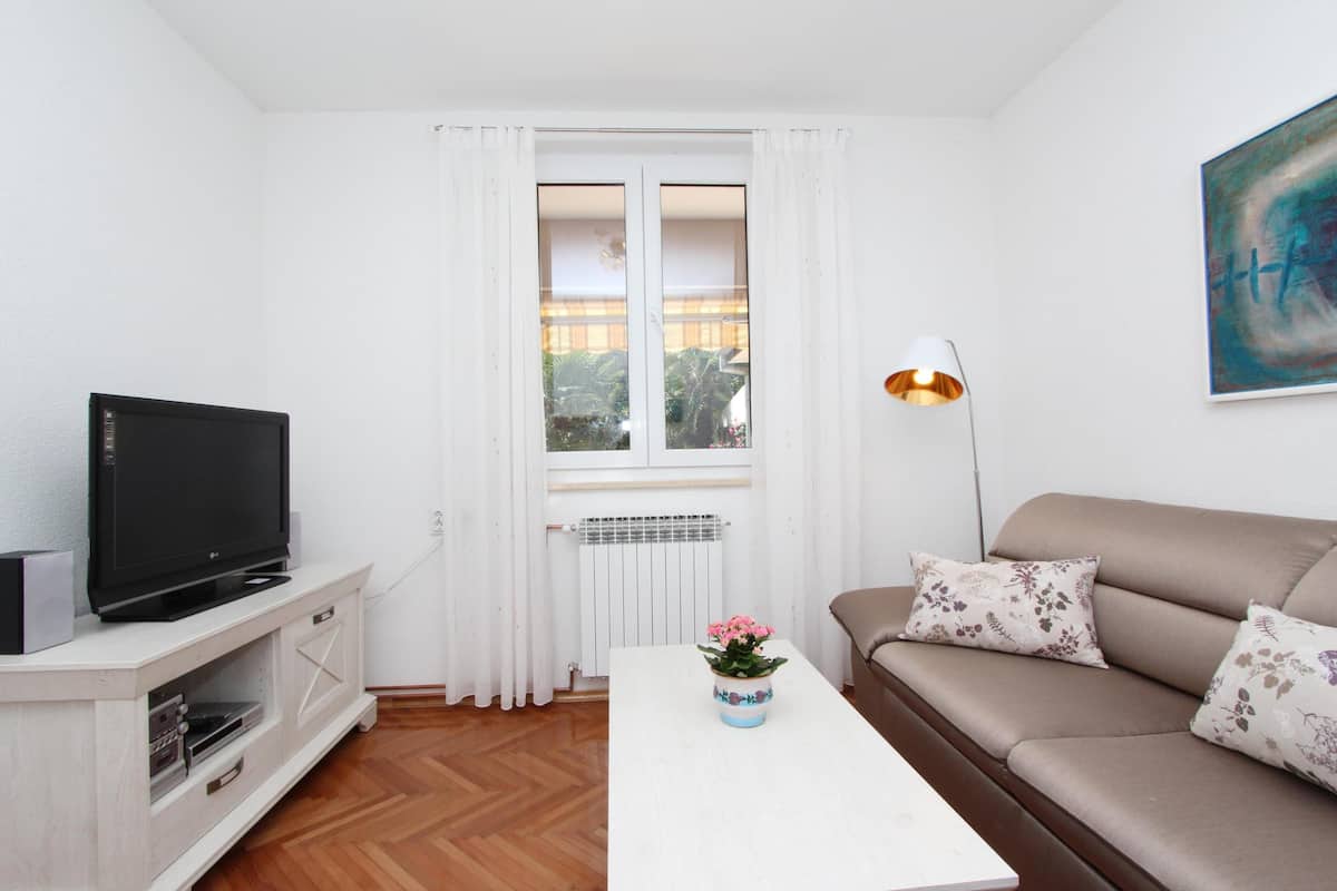Una sala de estar con una televisión sobre un mueble blanco, un sofá con cojines estampados, una mesa de centro blanca con un florero y una ventana con cortinas blancas.