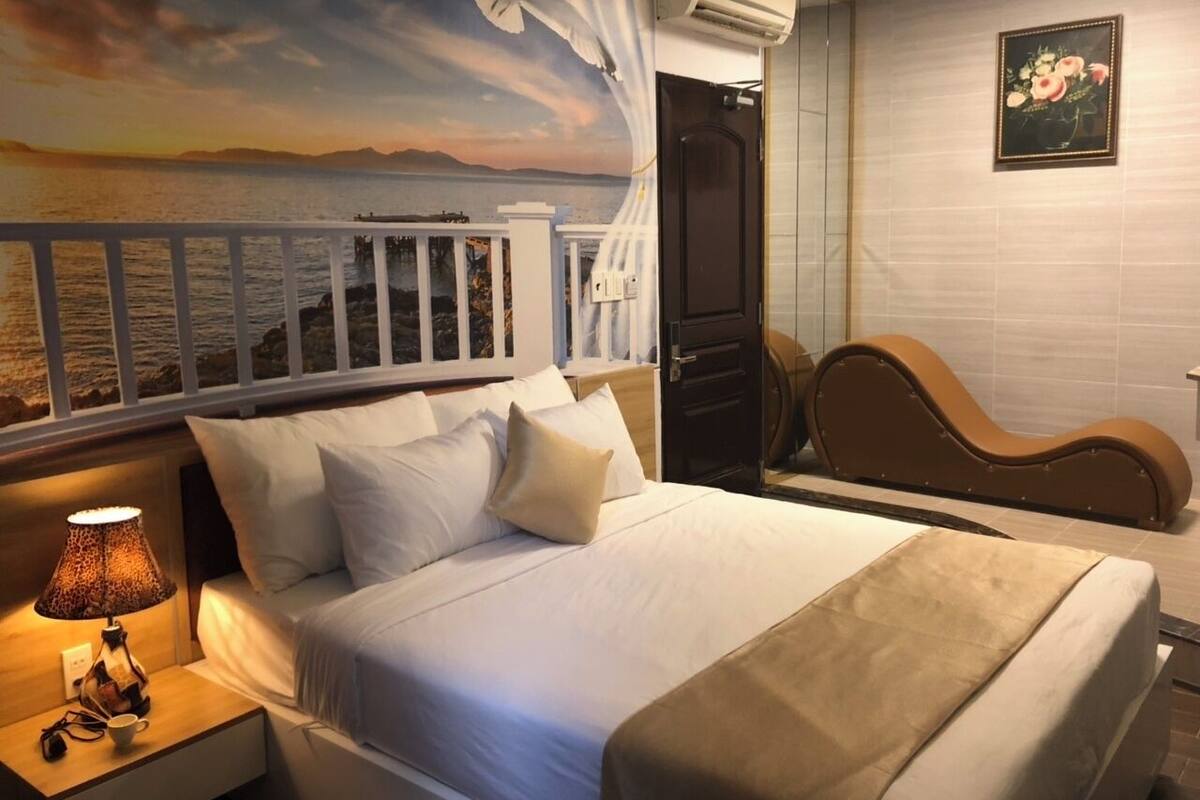 A hotel room with a bed, a nightstand with a lamp, a wooden headboard, and a painting of a sunset over the sea.