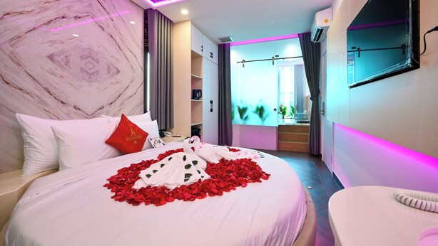 Una habitación de hotel moderna con pared de mármol, cama con pétalos de rosa, almohada roja y televisor en una esquina.
