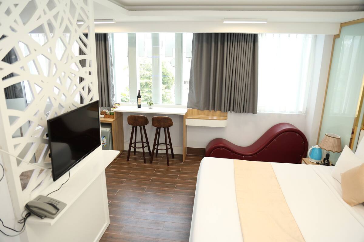 A hotel room with a bed, a TV, a desk with two stools, a window with curtains, and a mirror.
