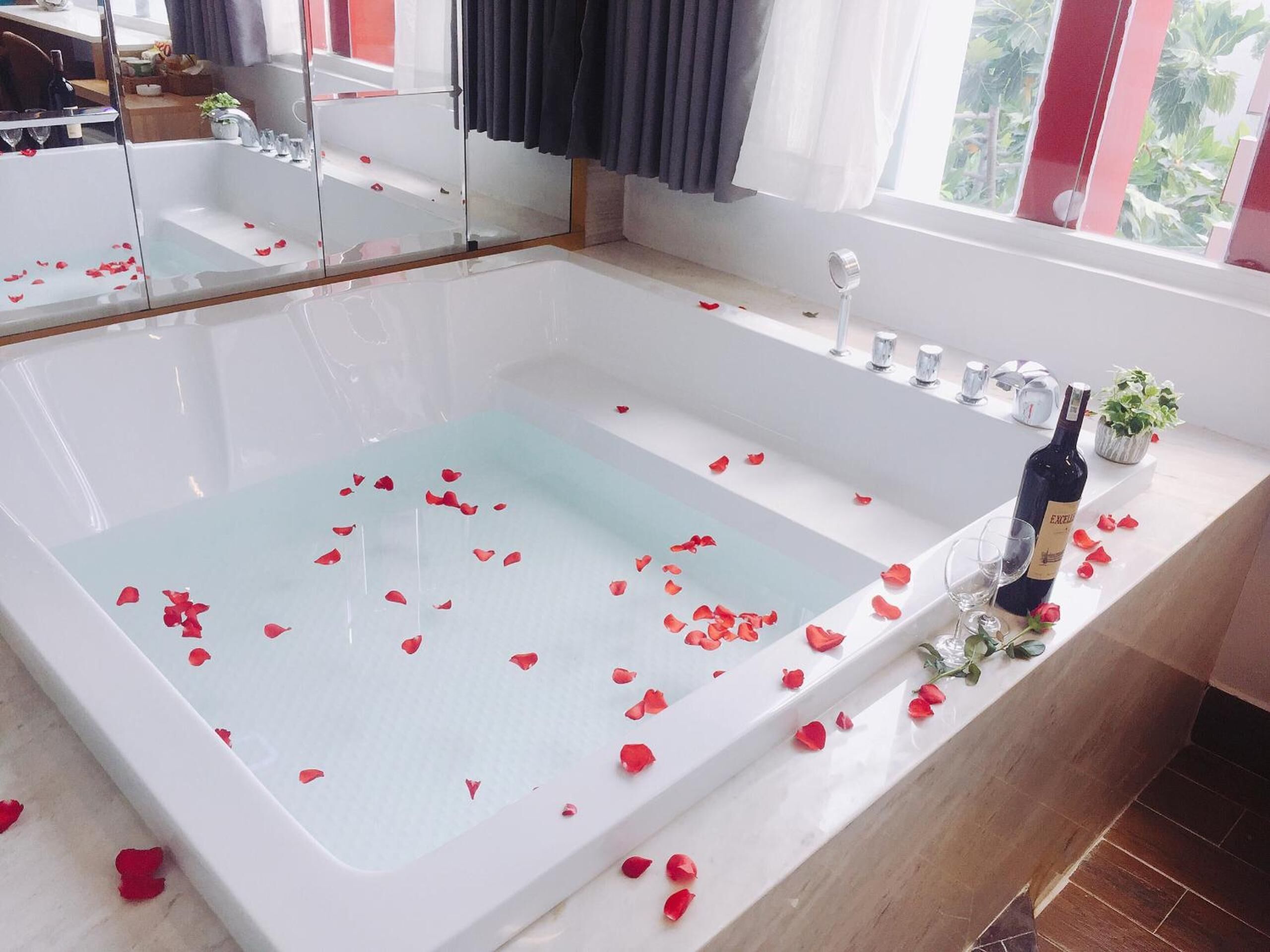 A modern bathroom with a large bathtub filled with rose petals, a wine bottle, and a small plant on the counter.