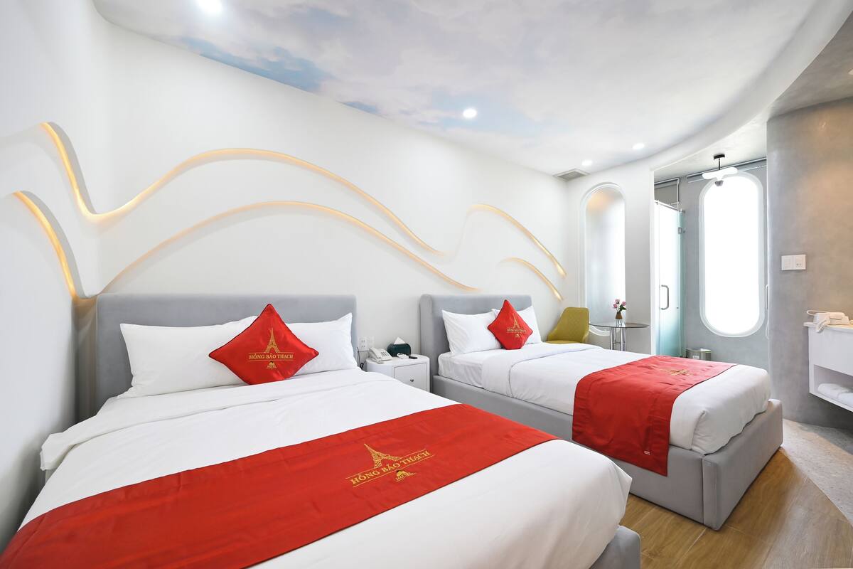 A hotel room with two beds, each with a red bedspread and white pillows. There is a small table with a lamp and a phone. The room has a modern design with a curved ceiling and a window with a view of the sky.
