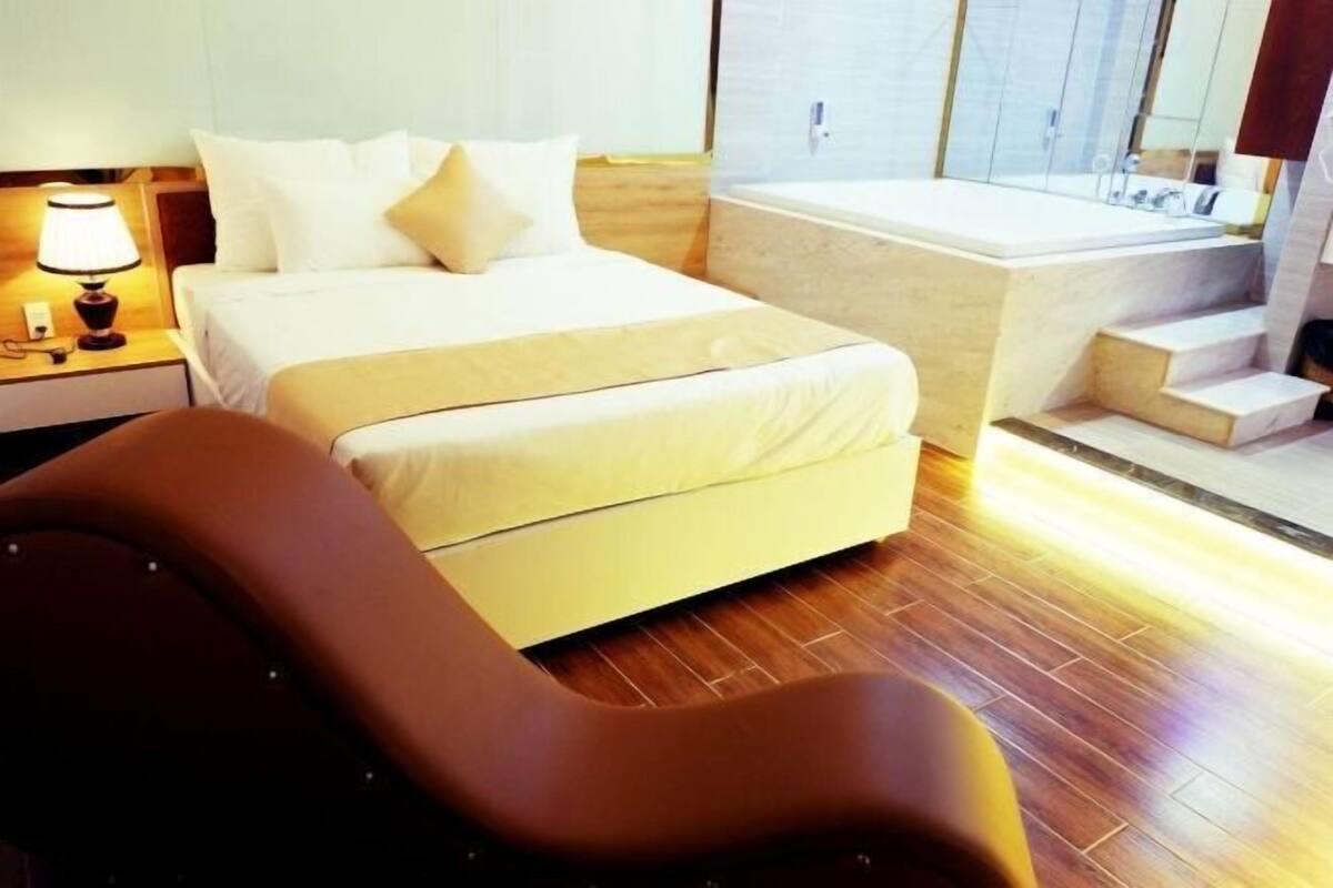 A hotel room with a bed, a nightstand with a lamp, a bathroom with a bathtub, and a wooden floor.