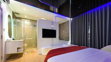 A modern hotel room with a large bed, a TV mounted on the wall, a shower area, and a vanity with a mirror.