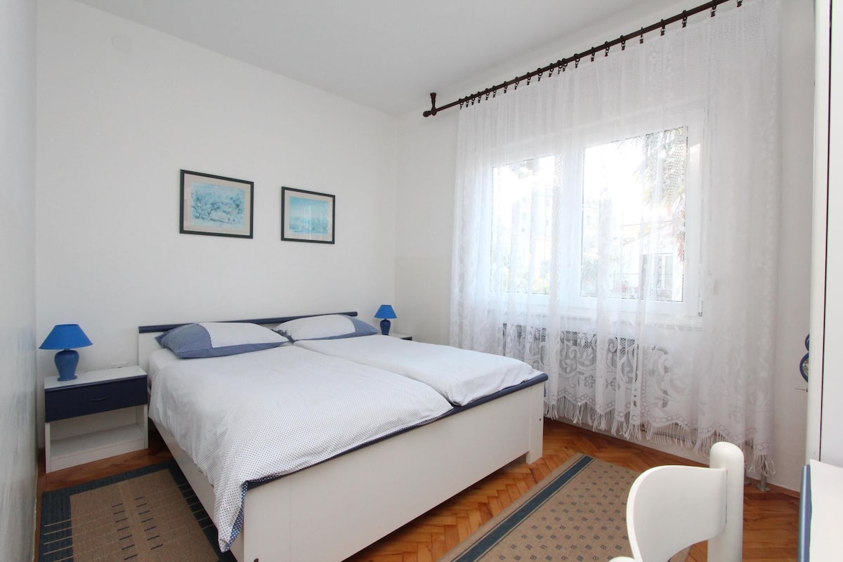 A bedroom with a bed, nightstands, a window with sheer curtains, and two framed pictures on the wall.