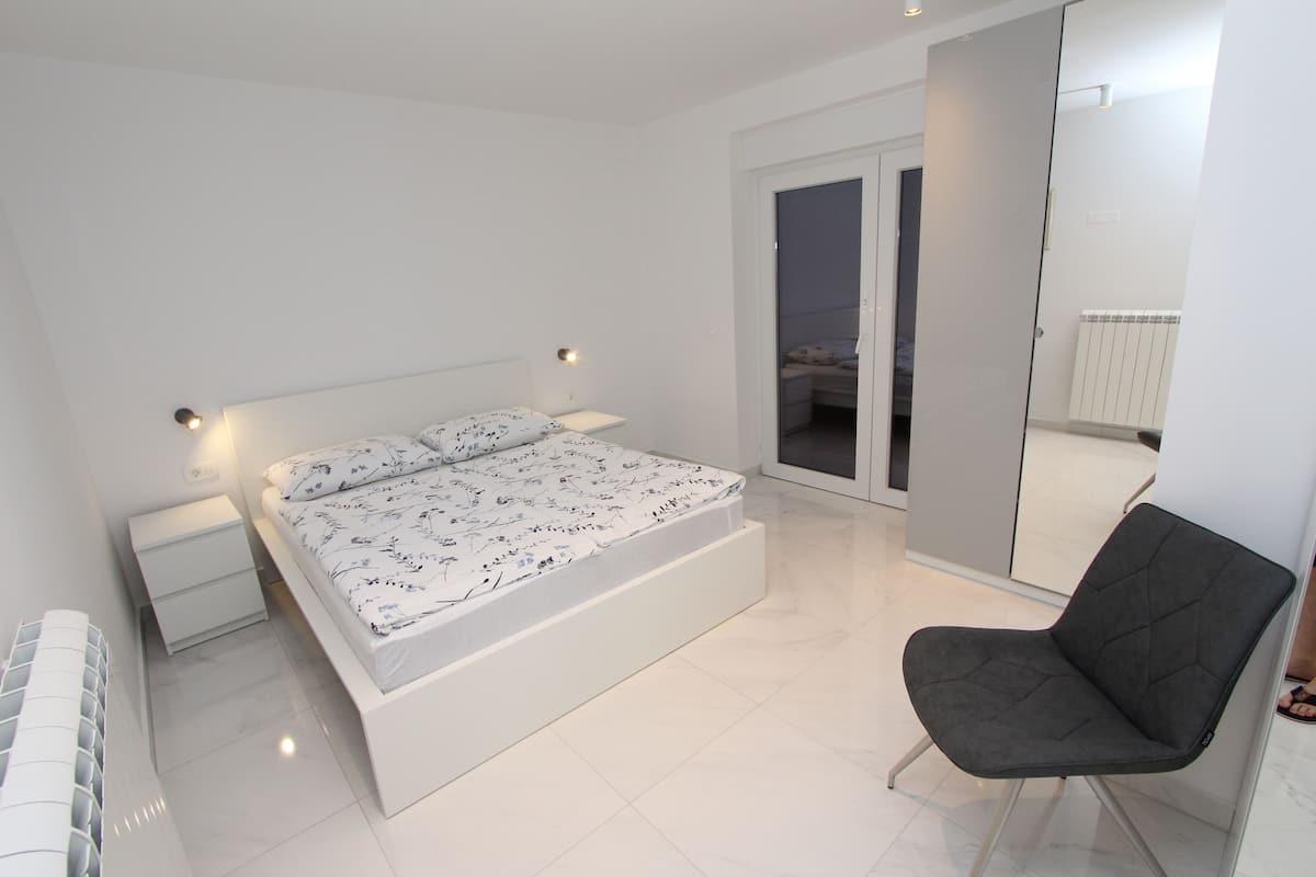 A modern bedroom with a bed, bedside tables, a chair, and a sliding glass door to a balcony.