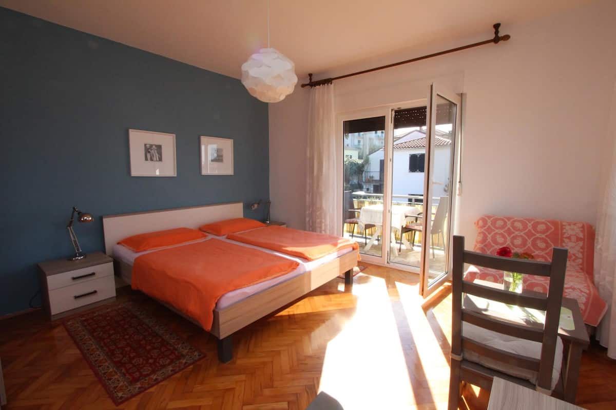 A bedroom with a double bed, a nightstand, a desk, a chair, and a view of the outside.