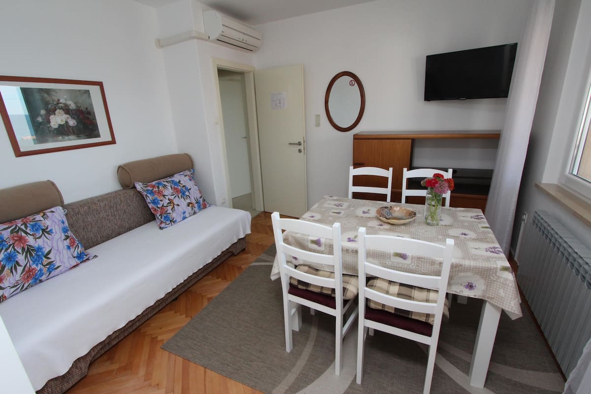 A room with a sofa, a dining table with chairs, a television mounted on the wall, and a radiator.