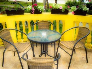 A round glass table surrounded by wicker chairs on a yellow balcony with potted plants.