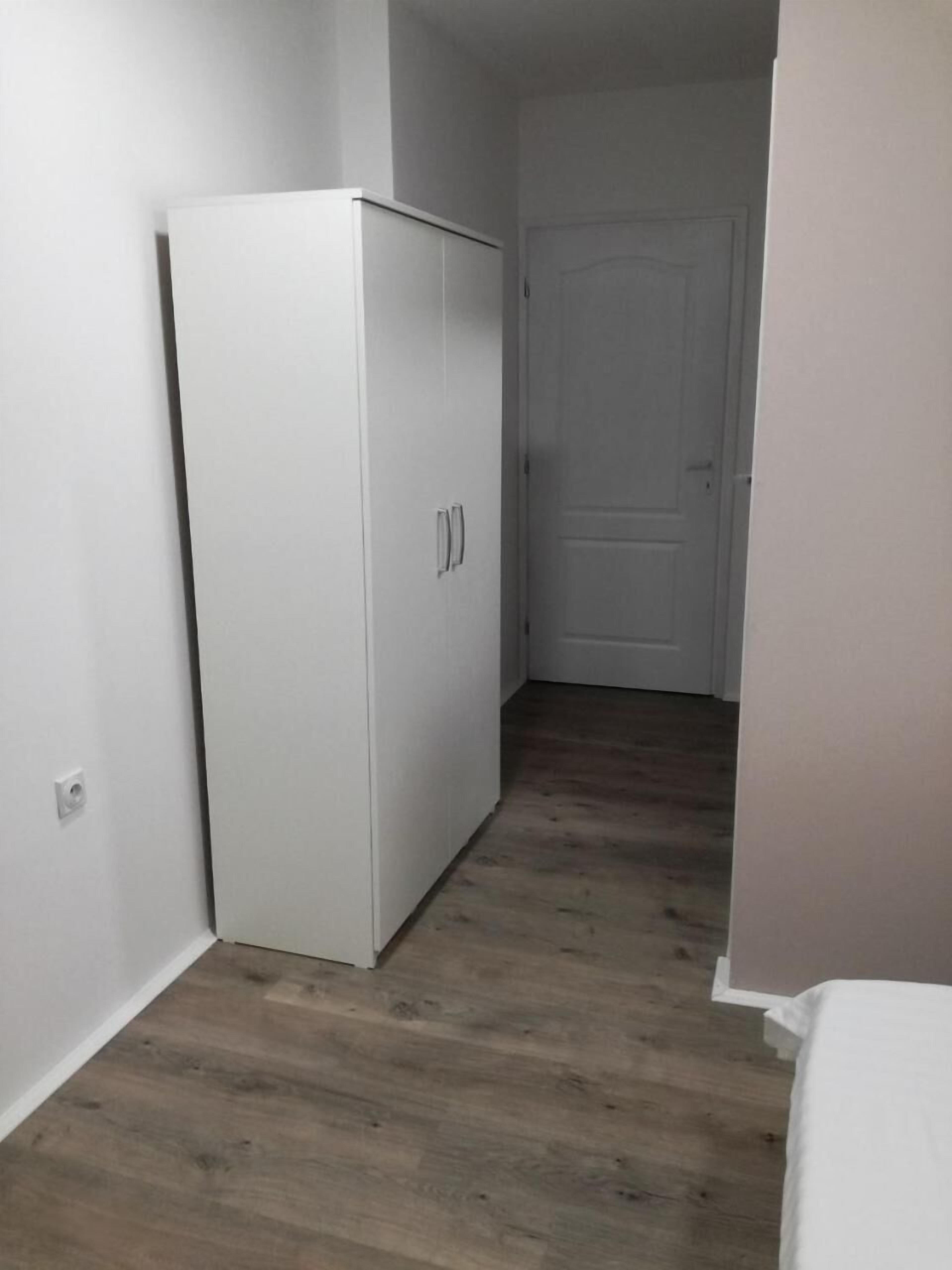 A room with a white wardrobe, a wooden floor, and a closed door in the background.