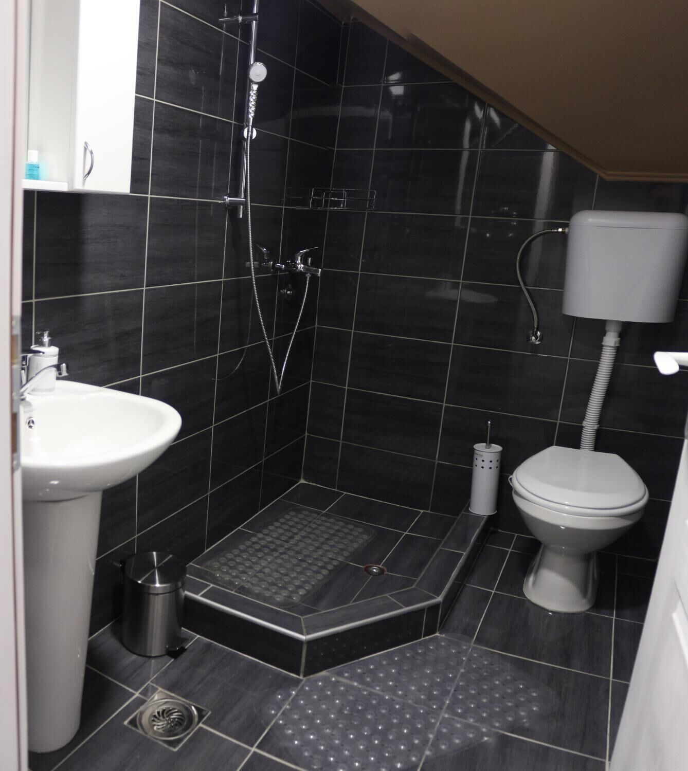Un bagno piccolo rivestito di piastrelle scure, con lavandino bianco, water e zona doccia.